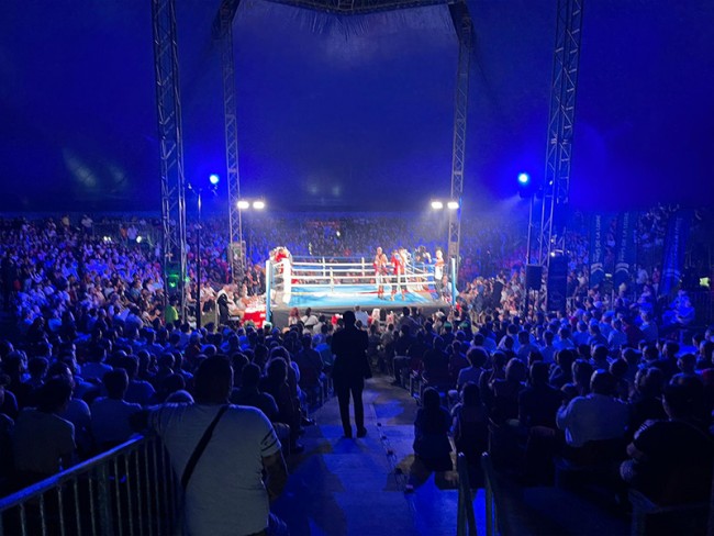 Photographie d'un spectacle de boxe, avec ring et gradins sous un chapiteau